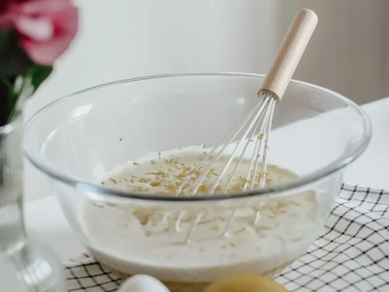 A whisk and baking ingredients in a glass mixing bowl, with eggs nearby.