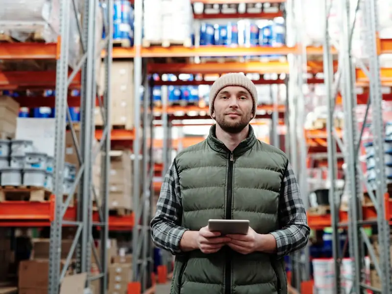 A person holding a tablet in an aisle of warehouse storage racks.
